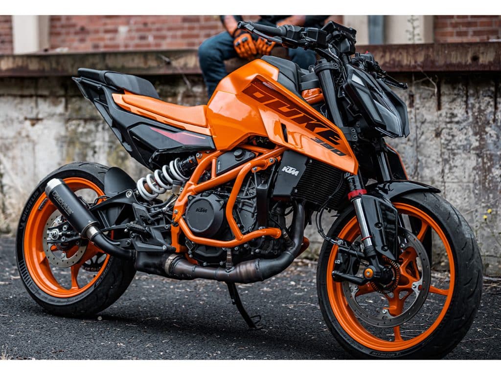 Test KTM 390 Duke A2 : garée en trois-quarts avant dans une ruelle urbaine, avec son pilote assis sur un muret en arrière-plan