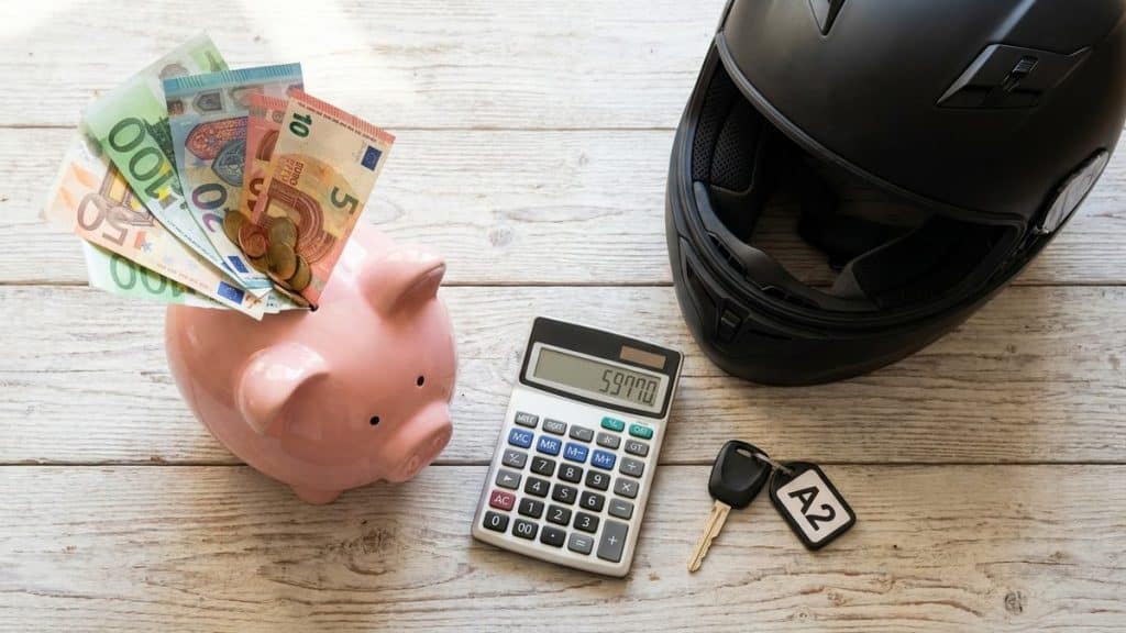 Flat-lay tirelire cochon rose billets euros calculatrice cle moto casque noir budget premiere A2